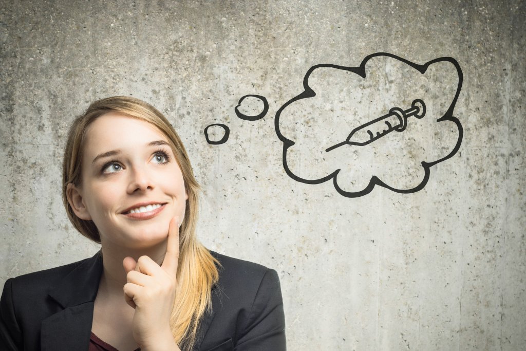 Eine junge Frau l&auml;chelt und schaut nachdenklich auf, den Finger am Kinn. Eine Gedankenblase mit der Zeichnung einer Spritze und dem Schriftzug "Impfen bei Apotheke Wyss" erscheint &uuml;ber ihrem Kopf vor einer strukturierten grauen Wand.