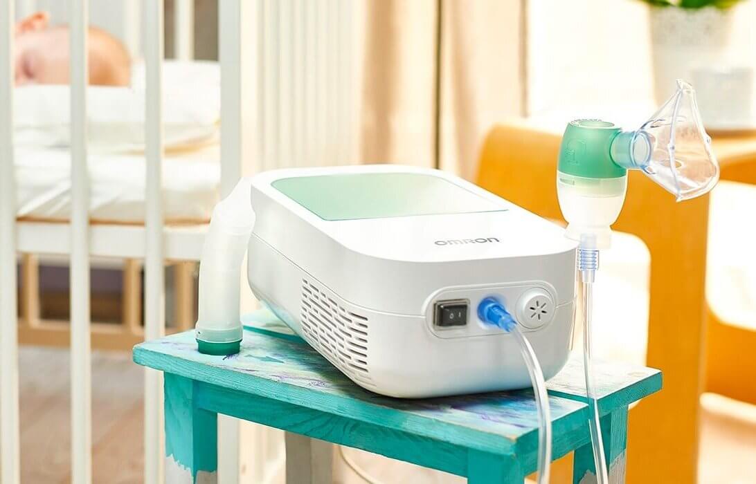 Ein Vernebelungsger&auml;t, ein unverzichtbarer Sanit&auml;tsartikel, steht auf einem kleinen, bunten Tisch in einem hellen Raum, mit einem Kinderbett und der verschwommenen Figur eines Babys, das auf einem Bett liegt, im Hintergrund. An dem Ger&auml;t sind eine Maske und ein Schlauch angebracht.