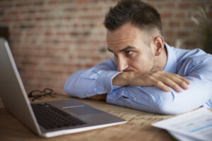 Ein Mann in einem blauen Hemd st&uuml;tzt seine Arme und sein Kinn auf einen Tisch. Er sieht m&uuml;de oder frustriert aus, w&auml;hrend er auf einen ge&ouml;ffneten Laptop starrt und sich m&ouml;glicherweise mit einem hartn&auml;ckigen Virus herumschl&auml;gt. Auf dem h&ouml;lzernen Schreibtisch liegen Gl&auml;ser und Papiere, im Hintergrund ist eine Backsteinmauer zu sehen.