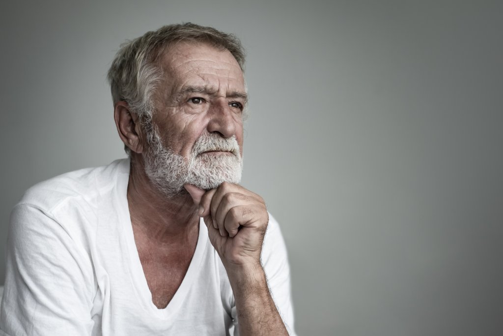 Ein &auml;lterer Mann mit grauem Haar und Bart, der ein wei&szlig;es T-Shirt tr&auml;gt, sitzt mit einer Hand, die nachdenklich auf seinem Kinn ruht, und blickt hartn&auml;ckig in die Ferne vor einem schlichten, hellgrauen Hintergrund.