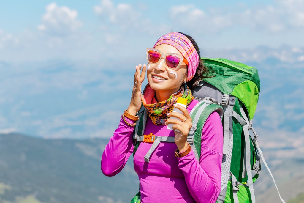 Eine l&auml;chelnde Frau in einem rosafarbenen Outfit und mit Sonnenbrille tr&auml;gt Sonnenschutz auf ihr Gesicht auf, w&auml;hrend sie in den Bergen wandert, einen gr&uuml;nen Rucksack und ein buntes Stirnband tr&auml;gt und eine malerische Landschaft im Hintergrund sieht.