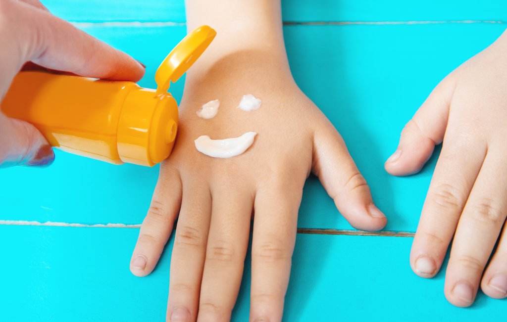 Eine Hand auf einer blauen Fl&auml;che mit Sonnenschutz, der aus einer gelben Flasche ausgepresst wird. Der Sonnenschutz bildet ein Smiley-Gesicht auf dem Handr&uuml;cken, w&auml;hrend eine andere Hand in der N&auml;he ruht.