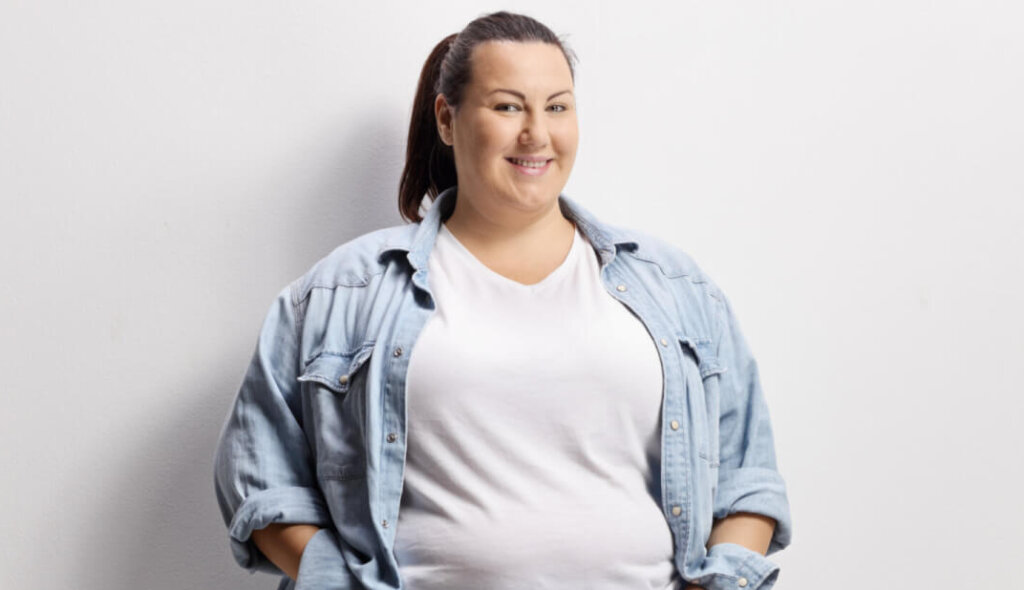 Eine Frau mit dunklem Haar in einem Pferdeschwanz, die ein helles Jeanshemd &uuml;ber einem wei&szlig;en T-Shirt tr&auml;gt, steht selbstbewusst l&auml;chelnd vor einem schlichten wei&szlig;en Hintergrund und verk&ouml;rpert Gleichgewicht und innere Ausgeglichenheit.