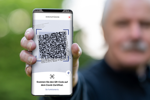 Eine Person h&auml;lt ein Smartphone in der Hand, auf dem ein gro&szlig;er QR-Code angezeigt wird, wahrscheinlich f&uuml;r ein COVID-Zertifikat, mit deutschem Text unter dem Code. Der Hintergrund ist unscharf und zeigt einen &auml;lteren Mann.