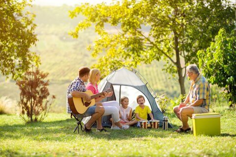 Eine Familie und ein &auml;lterer Mann sitzen mit ihrem Outdoorspass an einem Zelt im Freien. Der Mann spielt Gitarre, zwei Kinder spielen Schlagzeug, und alle l&auml;cheln und genie&szlig;en den sonnigen Tag inmitten von gr&uuml;nen B&auml;umen und Gras.