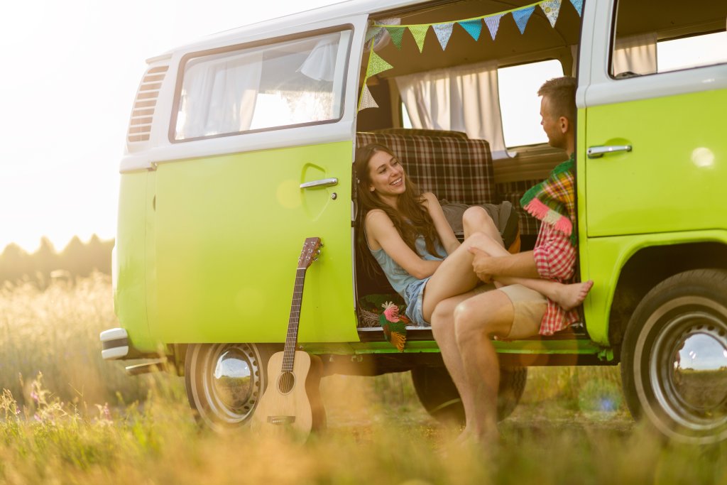 Ein junges Paar entspannt sich in einem gr&uuml;nen Retro-Van, der auf einer sonnigen Wiese geparkt ist, und genie&szlig;t sein Outdoorspass-Abenteuer. Die Frau lacht in der offenen T&uuml;r, w&auml;hrend der Mann drinnen sitzt, umgeben von einer Gitarre und Wimpeldekoration.