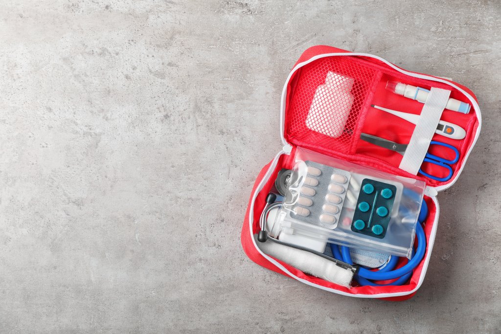 Ein roter Verbandskasten auf einer grauen Oberfl&auml;che, ideal f&uuml;r Outdoorspass-Abenteuer, enth&auml;lt Tabletten, eine Schere, eine Pinzette, einen Verband, Klebeband, Mull, eine kleine Flasche und ein blaues Stethoskop.