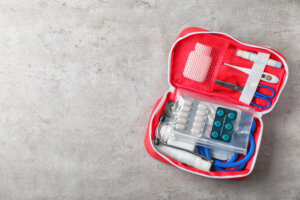 Ein roter Verbandskasten auf einer grauen Oberfl&auml;che, ideal f&uuml;r Outdoorspass-Abenteuer, enth&auml;lt Tabletten, eine Schere, eine Pinzette, einen Verband, Klebeband, Mull, eine kleine Flasche und ein blaues Stethoskop.