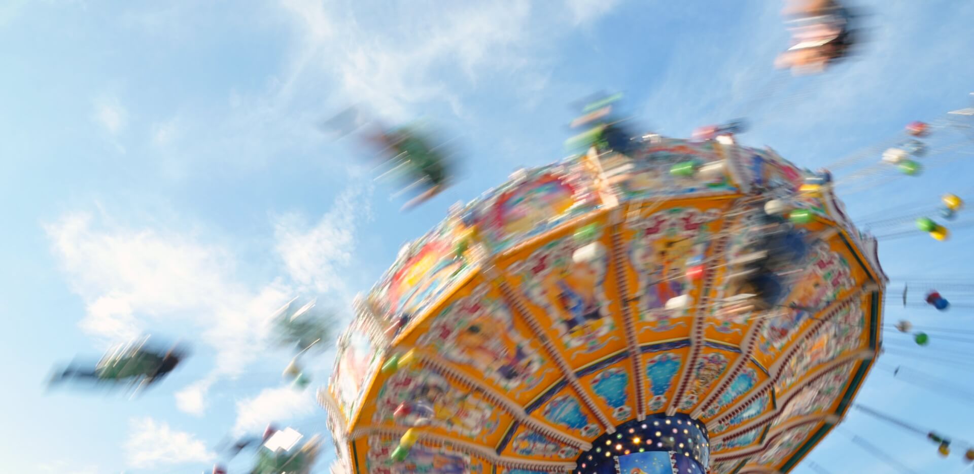 Eine farbenfrohe, sich drehende Karussellfahrt in einem Vergn&uuml;gungspark, aufgenommen mit Bewegungsunsch&auml;rfe vor einem blauen Himmel mit leichten Wolken, ruft Aufregung und den schwindelerregenden Rausch des Schwindels hervor.
