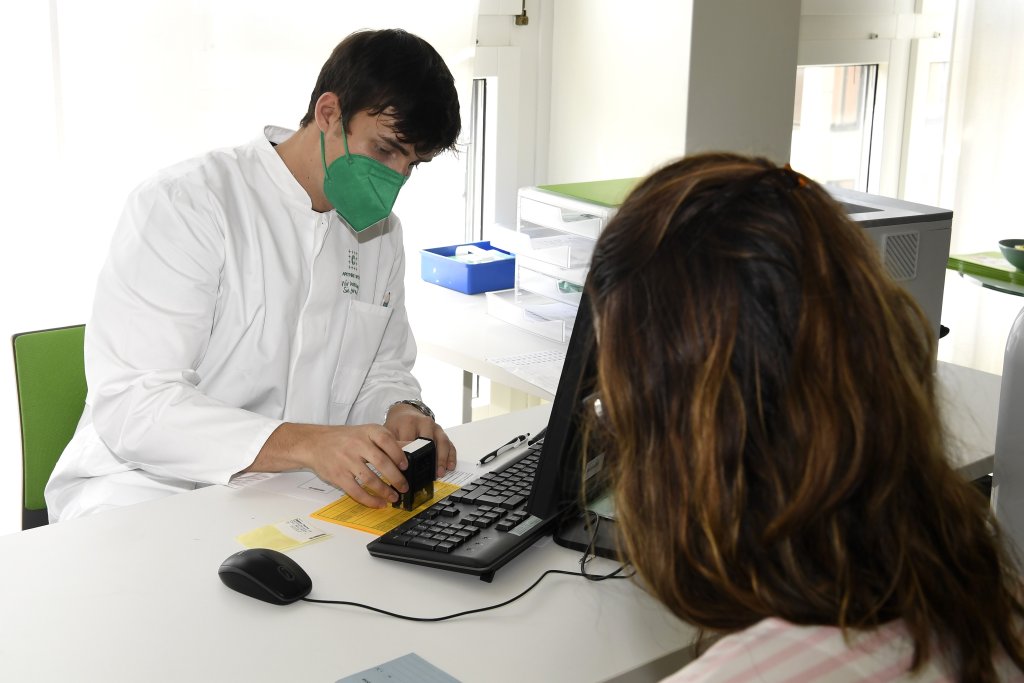 Ein DGS-Mitarbeiter in wei&szlig;em Kittel und gr&uuml;ner Maske stempelt w&auml;hrend eines Besuchs ein gelbes Dokument, w&auml;hrend ihm eine Frau mit braunem Haar teilweise von hinten sichtbar gegen&uuml;bersitzt.