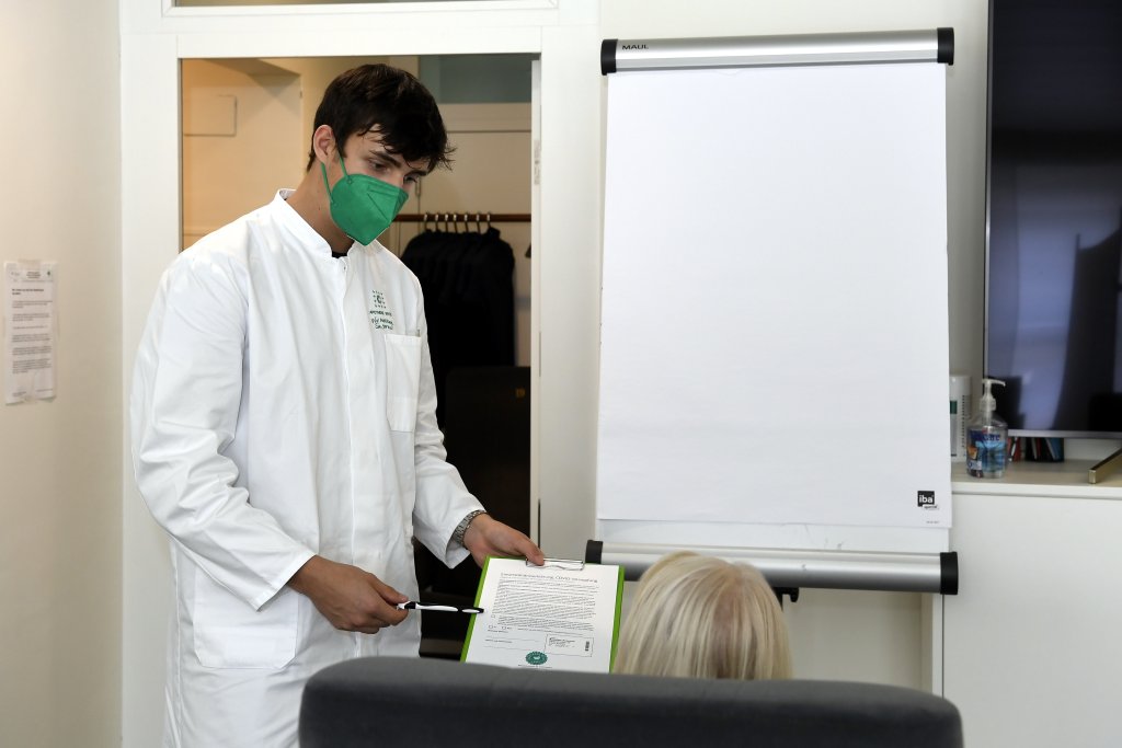 Ein Arzt mit wei&szlig;em Kittel und gr&uuml;ner Gesichtsmaske h&auml;lt ein Klemmbrett und einen Stift in der Hand, w&auml;hrend er einem sitzenden Patienten vor einem leeren Flipchart in einer Arztpraxis eine DGS-Visite erkl&auml;rt.