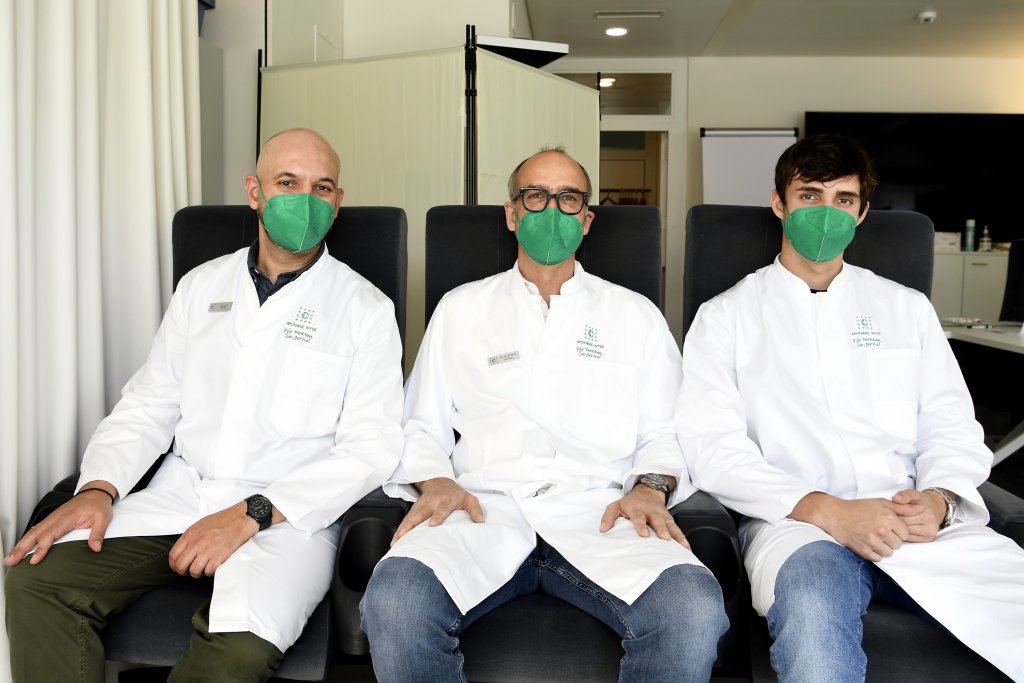 Drei M&auml;nner in wei&szlig;en Laborkitteln und gr&uuml;nen Gesichtsmasken sitzen w&auml;hrend eines DGS-Besuchs in einer klinischen Umgebung nebeneinander. Sie scheinen medizinische Fachkr&auml;fte zu sein, die auf dunklen St&uuml;hlen sitzen und in die Kamera schauen.