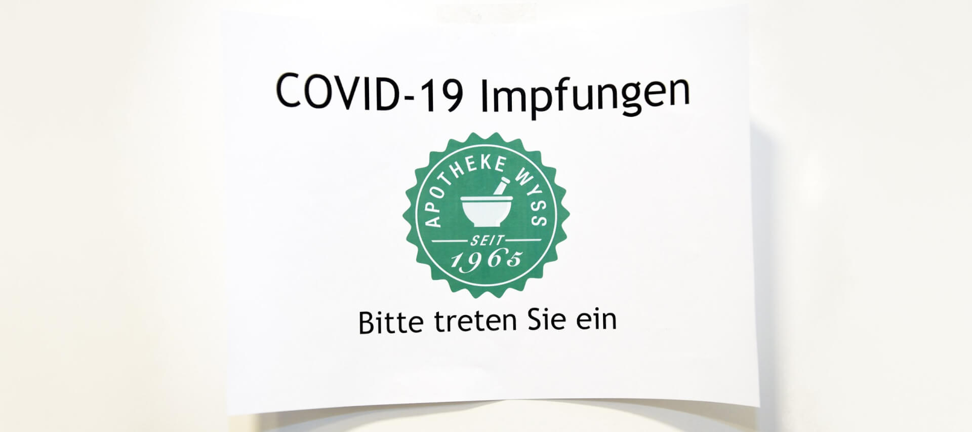 Ein Schild mit der Aufschrift COVID-19 Impfungen, darunter das Logo der Apotheke Wyss und der Text Bitte treten Sie ein und laden Ihren Besuch ein. DGS-Informationen k&ouml;nnen auch f&uuml;r die Zug&auml;nglichkeit verf&uuml;gbar sein.