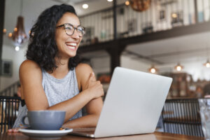Eine Frau mit lockigem Haar und Brille l&auml;chelt, w&auml;hrend sie mit einem Laptop und einer Kaffeetasse in einem hellen, modernen Caf&eacute; an einem Tisch sitzt.