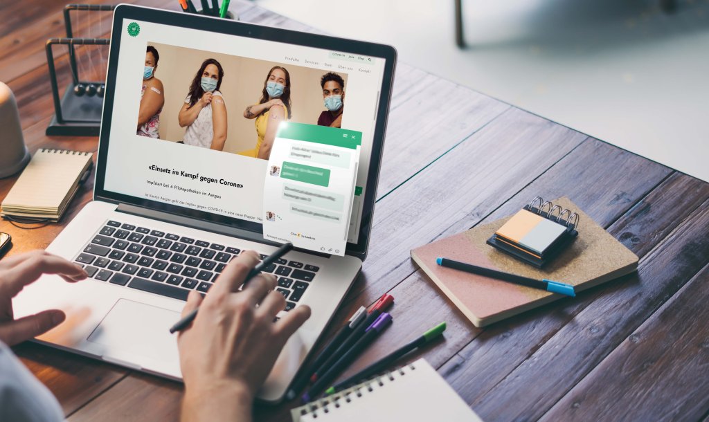 Eine Person arbeitet mit einem Laptop an einem Holztisch. Auf dem Bildschirm ist ein Bild von vier Personen mit Masken und hochgekrempelten &Auml;rmeln zu sehen, was auf eine Impfung hindeutet. Au&szlig;erdem sind ein Chat-Fenster, Stifte, Haftnotizen und eine Brille auf dem Schreibtisch zu sehen.