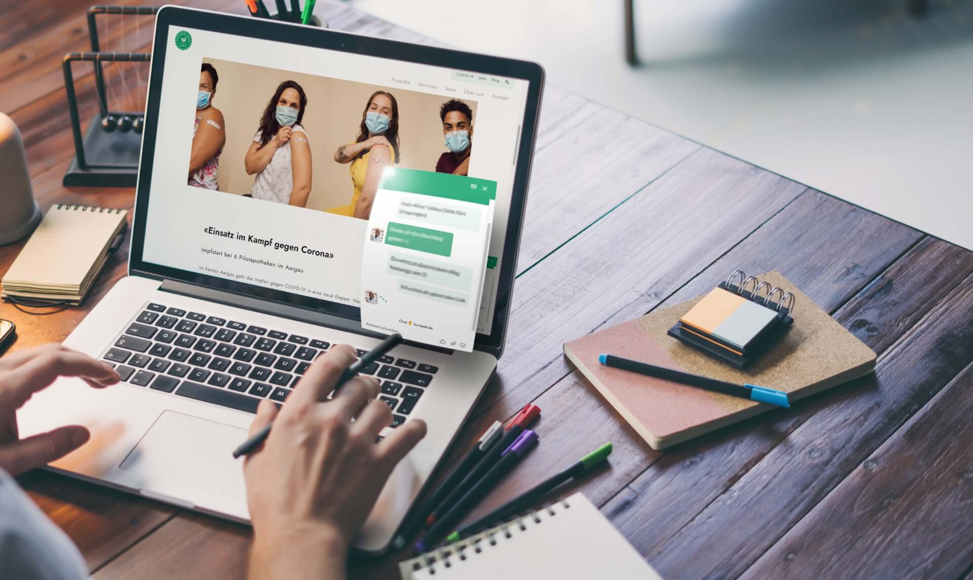 Eine Person arbeitet mit einem Laptop an einem Holztisch. Auf dem Bildschirm ist ein Bild von vier Personen mit Masken und hochgekrempelten &Auml;rmeln zu sehen, was auf eine Impfung hindeutet. Au&szlig;erdem sind ein Chat-Fenster, Stifte, Haftnotizen und eine Brille auf dem Schreibtisch zu sehen.
