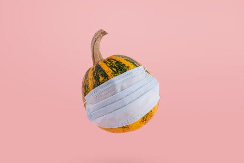 Ein kleiner, gr&uuml;n-gelb gestreifter K&uuml;rbis schwebt vor einem rosafarbenen Hintergrund und tr&auml;gt eine blaue medizinische Gesichtsmaske auf der Vorderseite - eine skurrile Anspielung auf die COVID-19-Sicherheit im Herbst 2021.