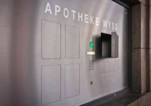 Ein metallisches Wandpaneel mit der Aufschrift APOTHEKE WYSS besteht aus mehreren rechteckigen Abschnitten, einer Tastatur, einem Display, einem gr&uuml;nen Aufkleber und einem offenen Fach - wahrscheinlich 24-Stunden-Abholf&auml;cher als Teil eines automatischen Apothekensystems.