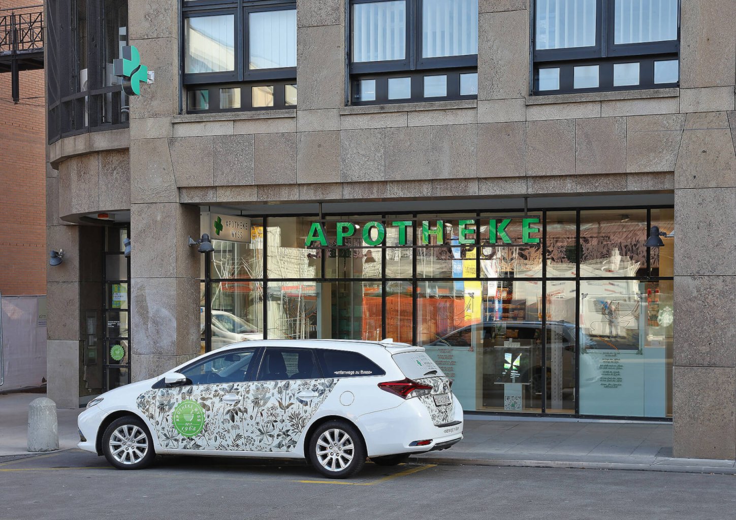 Ein wei&szlig;es Auto mit gr&uuml;ner Aufschrift parkt vor einer Apotheke, die 24-Stunden-Medikamenten-Abholf&auml;cher anbietet, mit gro&szlig;en Glasfenstern, einer Steinfassade und einem leuchtend gr&uuml;nen Apothekenschild &uuml;ber dem Eingang.