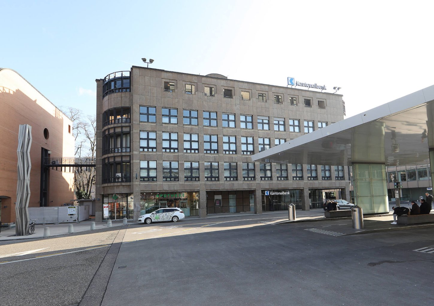 Ein modernes, mehrst&ouml;ckiges Geb&auml;ude mit gro&szlig;en Fenstern und einem Schild mit der Aufschrift "Stavanger tinghus" weist auf die nahe gelegenen 24h-Medikamenten-Abholf&auml;cher hin. Im Vordergrund befinden sich einige geparkte Autos, ein &uuml;berdachter Gehweg und Menschen, die auf B&auml;nken sitzen.