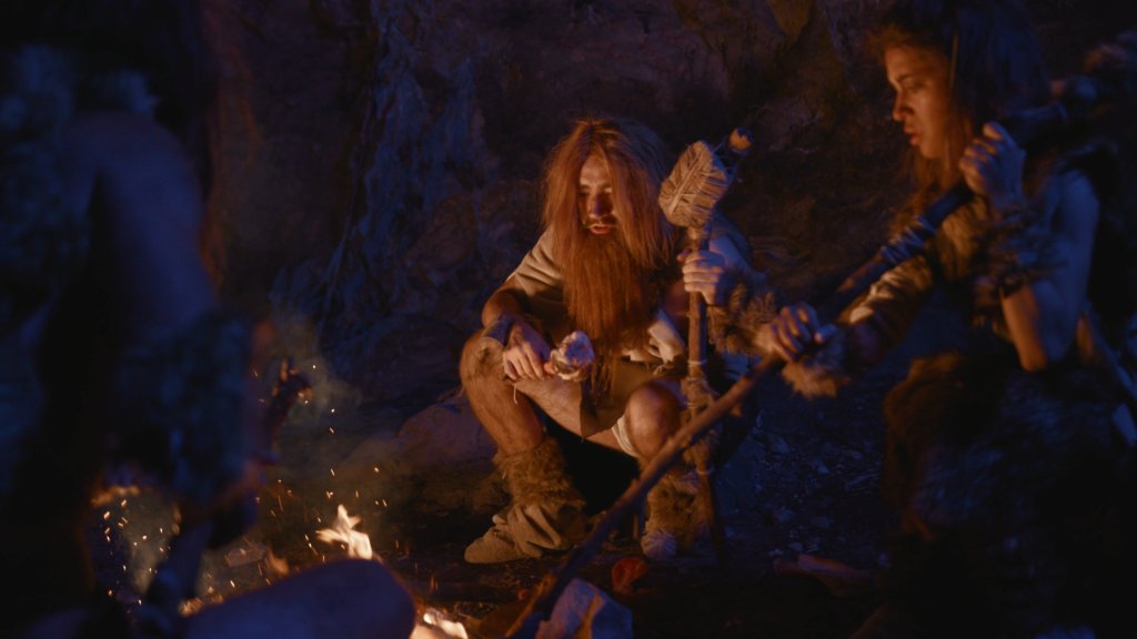 Drei als pr&auml;historische Menschen verkleidete Personen sitzen in einer schwach beleuchteten H&ouml;hle um ein Lagerfeuer, halten primitive Werkzeuge und St&ouml;cke in der Hand und entdecken die faszinierende Welt der Pilze im warmen Feuerschein.