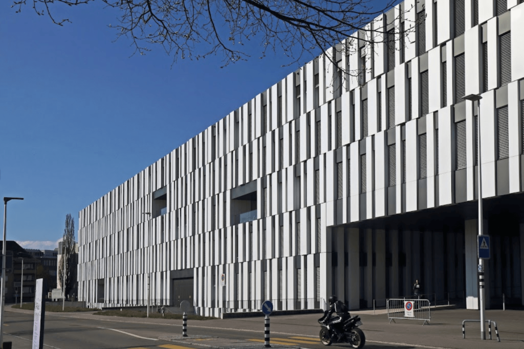 Ein modernes wei&szlig;es Geb&auml;ude mit vertikalen Linien und rechteckigen Fenstern steht am Rande einer Stra&szlig;e. Eine Person auf einem Motorrad, die vielleicht &uuml;ber Medikation im Alter nachdenkt, f&auml;hrt unter blauem Himmel und kahlen Baumzweigen vorbei.