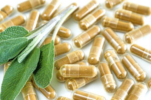Mehrere gr&uuml;ne Bl&auml;tter ruhen auf einer Gruppe von braunen, durchsichtigen Tee-Tablettenkapseln, die pulverisierte pflanzliche Nahrungserg&auml;nzungsmittel auf einer wei&szlig;en Oberfl&auml;che enthalten.