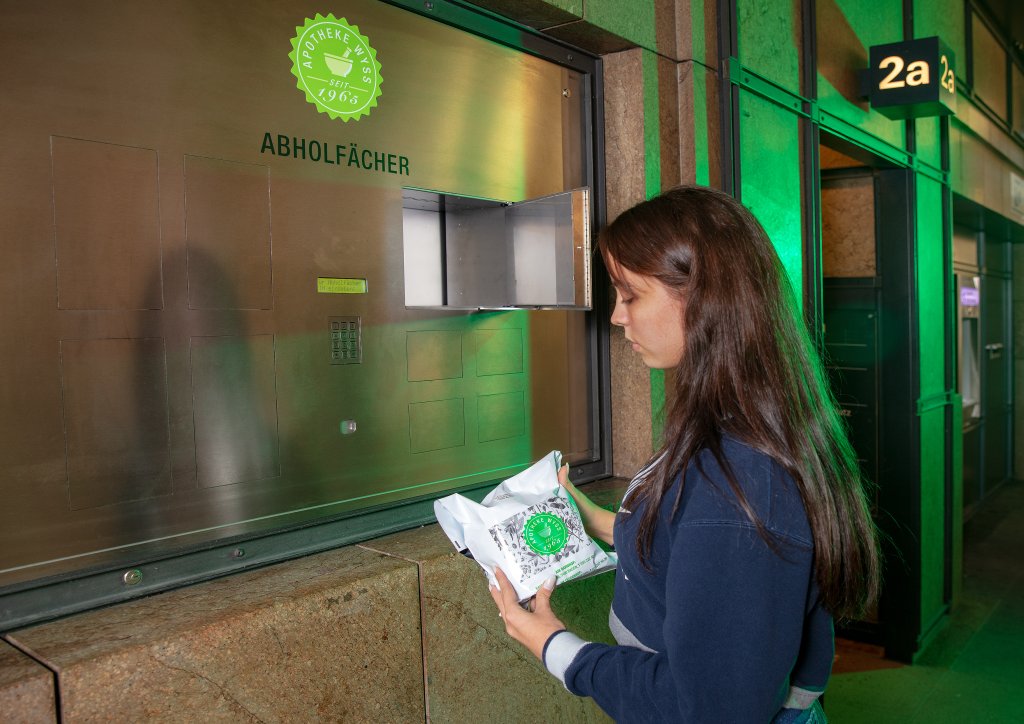 Eine Frau steht an einem 24-Std.-Abholf&auml;cher aus Metall und h&auml;lt ein wei&szlig;es Paket mit einem gr&uuml;nen Siegel in der Hand, das sie in ein offenes Fach einlegen oder herausnehmen will. Der Schauplatz scheint eine Inneneinrichtung zu sein.