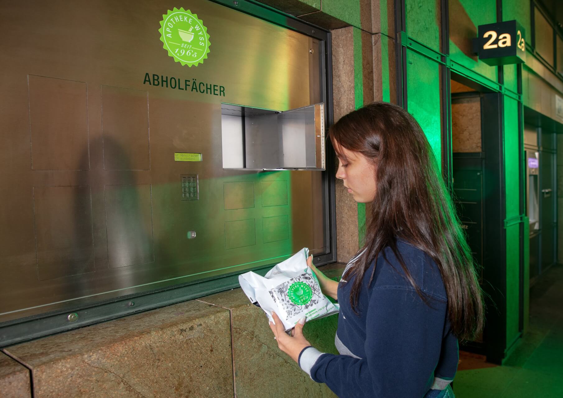 Eine Frau steht an einem 24-Std.-Abholf&auml;cher aus Metall und h&auml;lt ein wei&szlig;es Paket mit einem gr&uuml;nen Siegel in der Hand, das sie in ein offenes Fach einlegen oder herausnehmen will. Der Schauplatz scheint eine Inneneinrichtung zu sein.