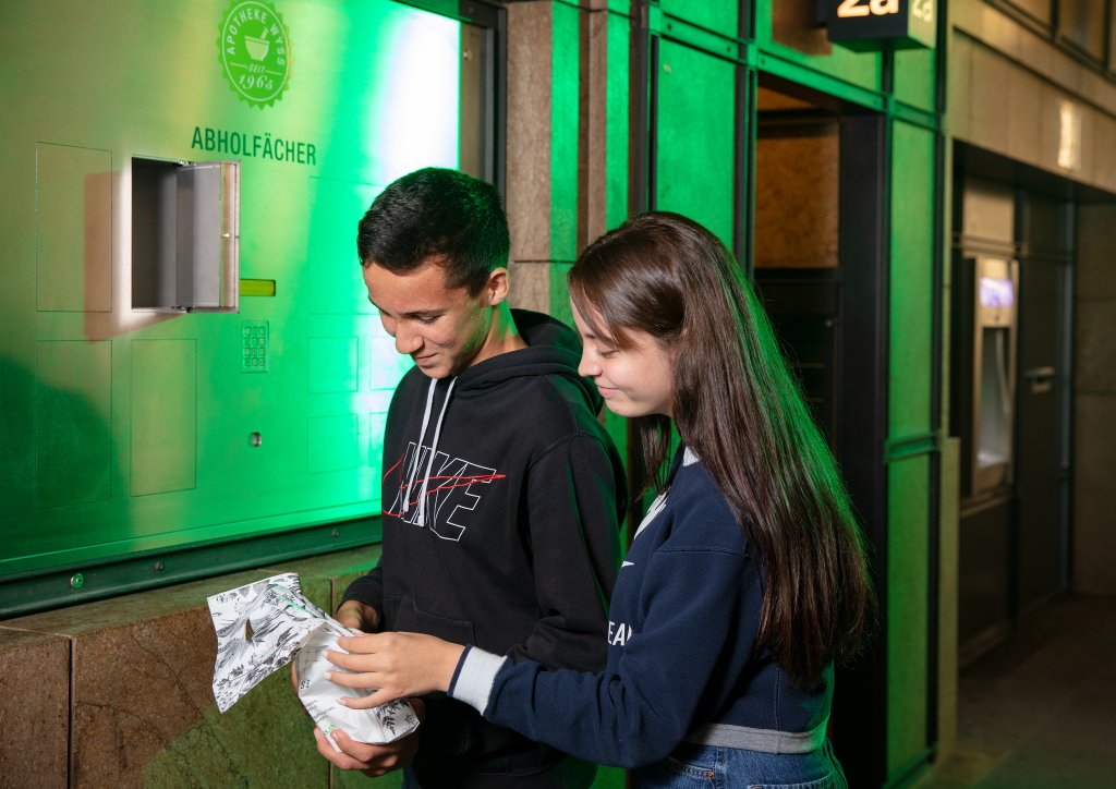 Zwei Teenager stehen an einem Wandschrank mit einem kleinen offenen Fach-24-Std-Abholf&auml;cher. Der Junge tr&auml;gt einen schwarzen Nike-Kapuzenpulli, und das M&auml;dchen steht neben ihm, w&auml;hrend sie gemeinsam unter gr&uuml;nem Licht ein wei&szlig;es Papierpaket &ouml;ffnen.