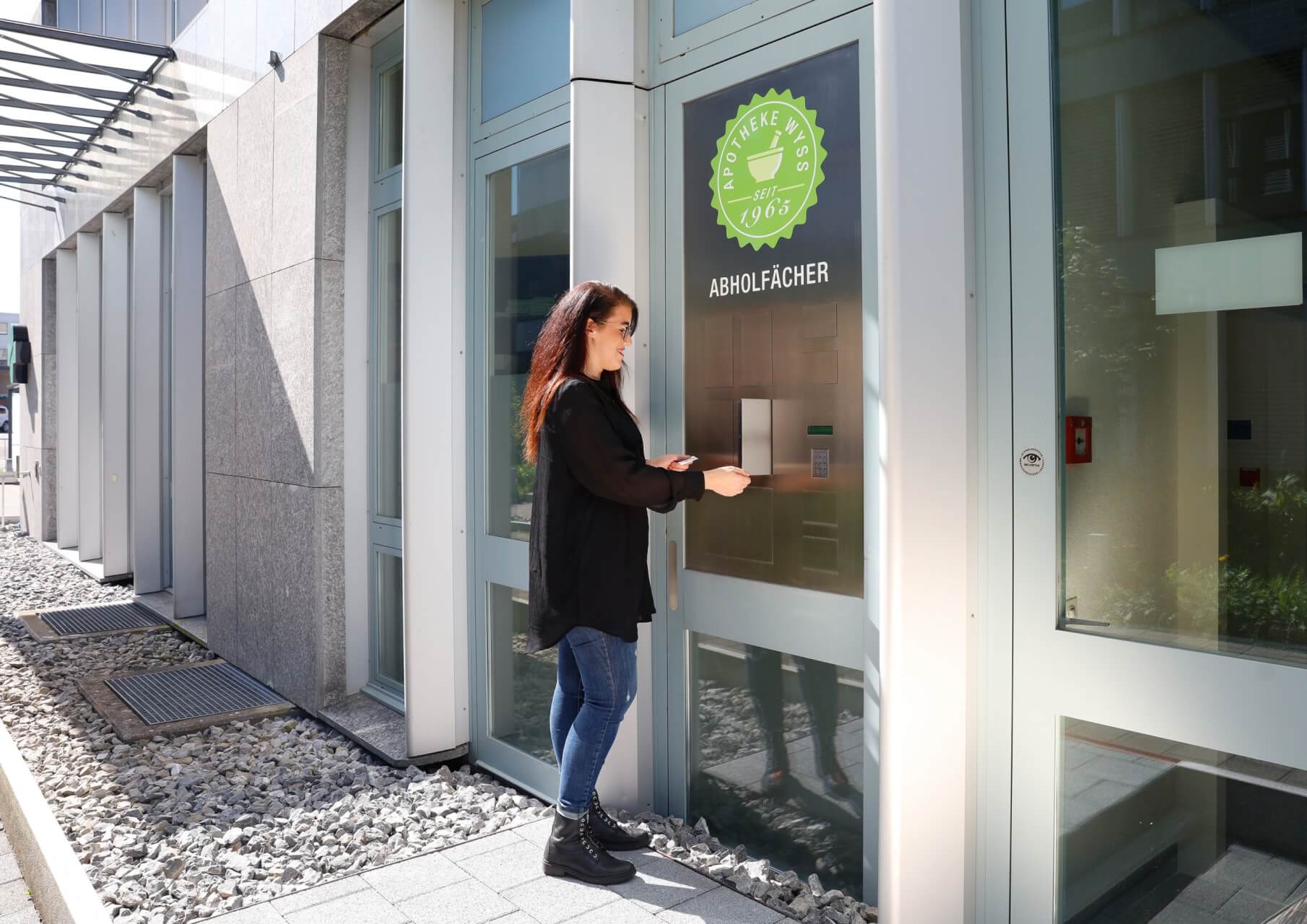 Eine Frau steht vor einem modernen Geb&auml;ude und benutzt ein an der Wand befestigtes Schlie&szlig;fach mit einem gr&uuml;nen Aufkleber mit der Aufschrift 24-Std-Abholf&auml;cher, was auf Deutsch 24-Stunden-Abholf&auml;cher bedeutet. Sie holt einen Gegenstand aus einem Sicherheitsfach.