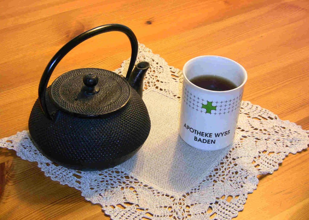 Eine schwarze gusseiserne Teekanne und eine wei&szlig;e Tasse mit der Aufschrift "APOTHEKE WYSS BADEN" stehen auf einem Spitzendeckchen auf einem Holztisch. In der Tasse befindet sich Tee, den man in dieser gem&uuml;tlichen Umgebung genie&szlig;en kann.