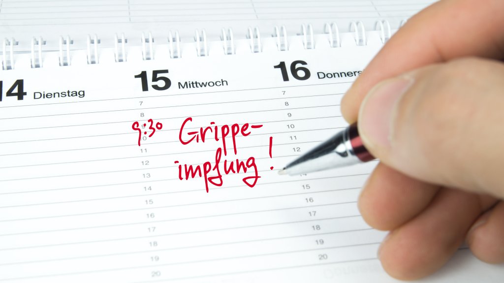 Eine Hand schreibt mit roter Tinte "9:30 Grippeimpfung 2022!" auf einen deutschen Kalender, der auf Mittwoch, den 15. Januar, aufgeschlagen ist, und h&auml;lt einen Stift in die N&auml;he der handschriftlichen Notiz.