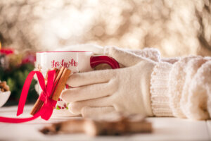 Eine Person mit wei&szlig;en Strickhandschuhen h&auml;lt einen festlichen Adventsbecher mit der Aufschrift Ho Ho Ho, verziert mit einem roten Band und Zimtstangen, vor einem weichen, warmen und verschwommenen Winterhintergrund.