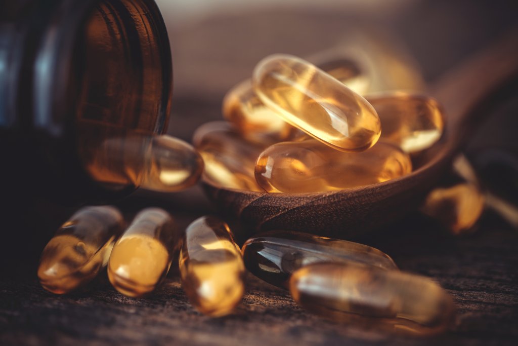 Eine Nahaufnahme mehrerer durchsichtiger, goldener Vitamin-D-Kapseln, die oft als Sonnenvitamin bezeichnet werden, aus einer Flasche, von denen einige auf einem Holzl&ouml;ffel ruhen und andere auf einer rustikalen Holzoberfl&auml;che verstreut sind.