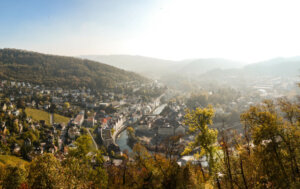 Der Panoramablick auf eine Stadt, die in einem Tal liegt, durch das sich ein Fluss schl&auml;ngelt, umgeben von bewaldeten H&uuml;geln und verstreuten H&auml;usern unter einem dunstigen, sonnenbeschienenen Himmel, erinnert an die ruhige Resilienz in der &Auml;ra von COVID-19.