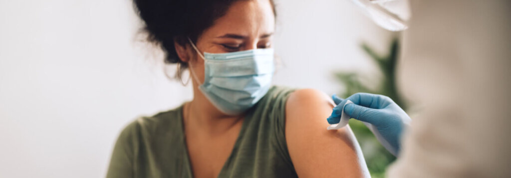 Eine Frau mit Gesichtsmaske schaut weg, w&auml;hrend eine medizinische Fachkraft mit Handschuhen ihren Oberarm mit einem Wattebausch s&auml;ubert, um sich auf eine COVID-19-Auffrischungsimpfung vorzubereiten.
