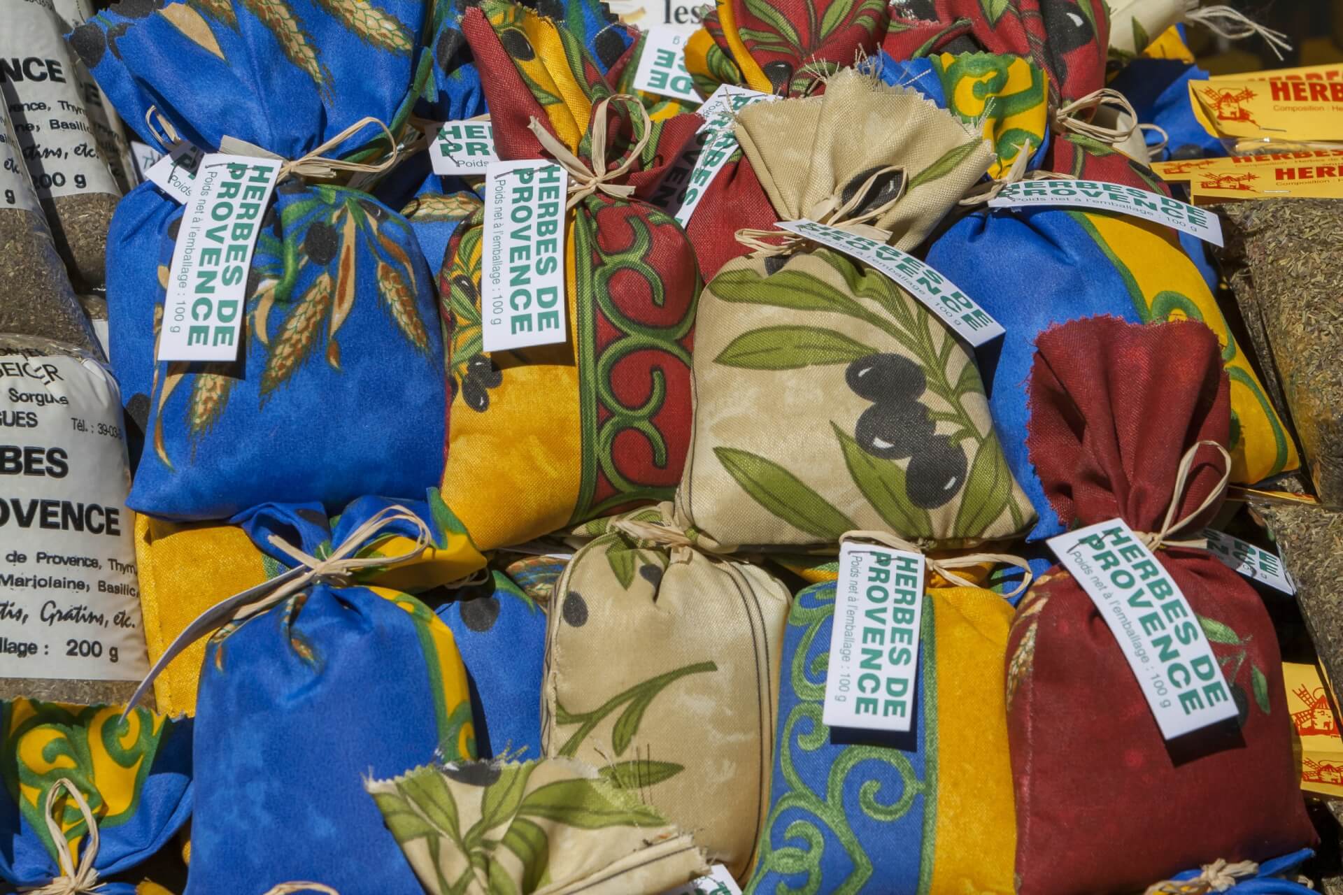 Bunte Stoffbeutel, gef&uuml;llt mit Herbes de Provence und anderen Gew&uuml;rzen als Heilmittel, sind &uuml;bereinander gestapelt, jeder mit einem Etikett versehen. Die Beutel zeigen Muster wie Oliven und Bl&auml;tter als Teil einer einladenden Auslage f&uuml;r einen Markt im Freien.