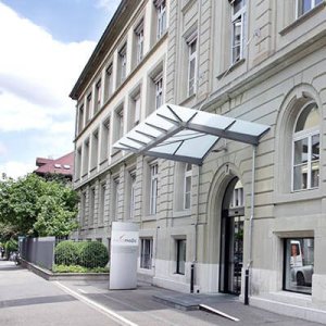 Eingang zu einem beigen Steingeb&auml;ude mit gro&szlig;en Bogenfenstern und einem modernen Glasvordach &uuml;ber dem Eingang; ein Schild mit einem Logo und der Aktualisierung COVID-19 steht in der N&auml;he des Eingangs entlang eines Gehwegs mit B&auml;umen und Gr&uuml;nfl&auml;chen.