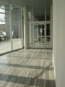Ein sonnendurchfluteter Korridor mit grossen Glasfenstern und einer Glast&uuml;r am Ende im T&auml;fernhof D&auml;ttwil. Der grau marmorierte Boden f&auml;ngt Sonnenlicht und Schatten ein, w&auml;hrend draussen ein Teil eines Autos und Geb&auml;ude das 15-j&auml;hrige Jubil&auml;um markieren.