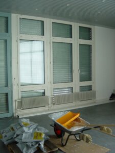 Ein heller Raum mit grossen, mit Jalousien abgedeckten Fenstern im T&auml;fernhof D&auml;ttwil, darunter ein Heizk&ouml;rper und auf dem Boden Baumaterialien wie eine Schubkarre, Sands&auml;cke und Werkzeuge - so wird der Geist von 15 Jahre Jubil&auml;um eingefangen.