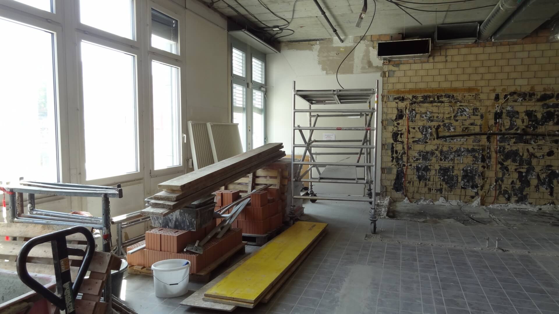 Ein unfertiger Raum im T&auml;fernhof D&auml;ttwil, der gerade renoviert wird. Ger&uuml;ste, Holzbretter, Ziegelsteine und Werkzeuge liegen auf dem gefliesten Boden verstreut; freiliegende Ziegel und Dr&auml;hte deuten auf 15 Jahre handwerklicher Tradition hin.