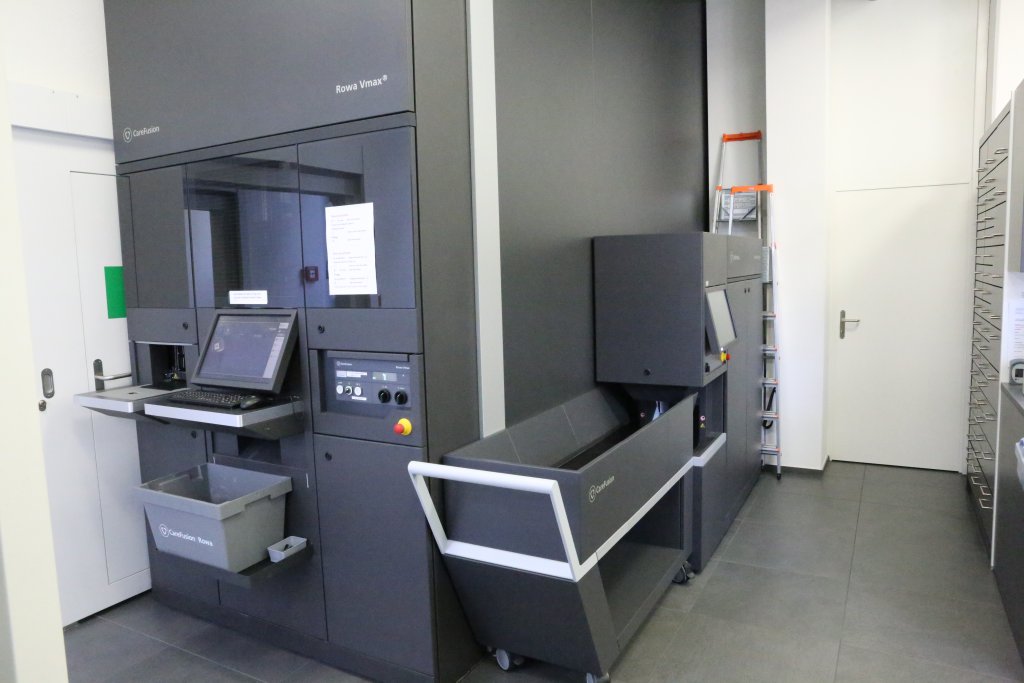 Eine moderne Buchsortiermaschine mit Computerterminal, grauen Beh&auml;ltern und einer Leiter im Hintergrund steht in einem sauberen, weissen Raum des T&auml;fernhofs D&auml;ttwil, der mit effizienter Organisation 15 Jahre Jubil&auml;um feiert.