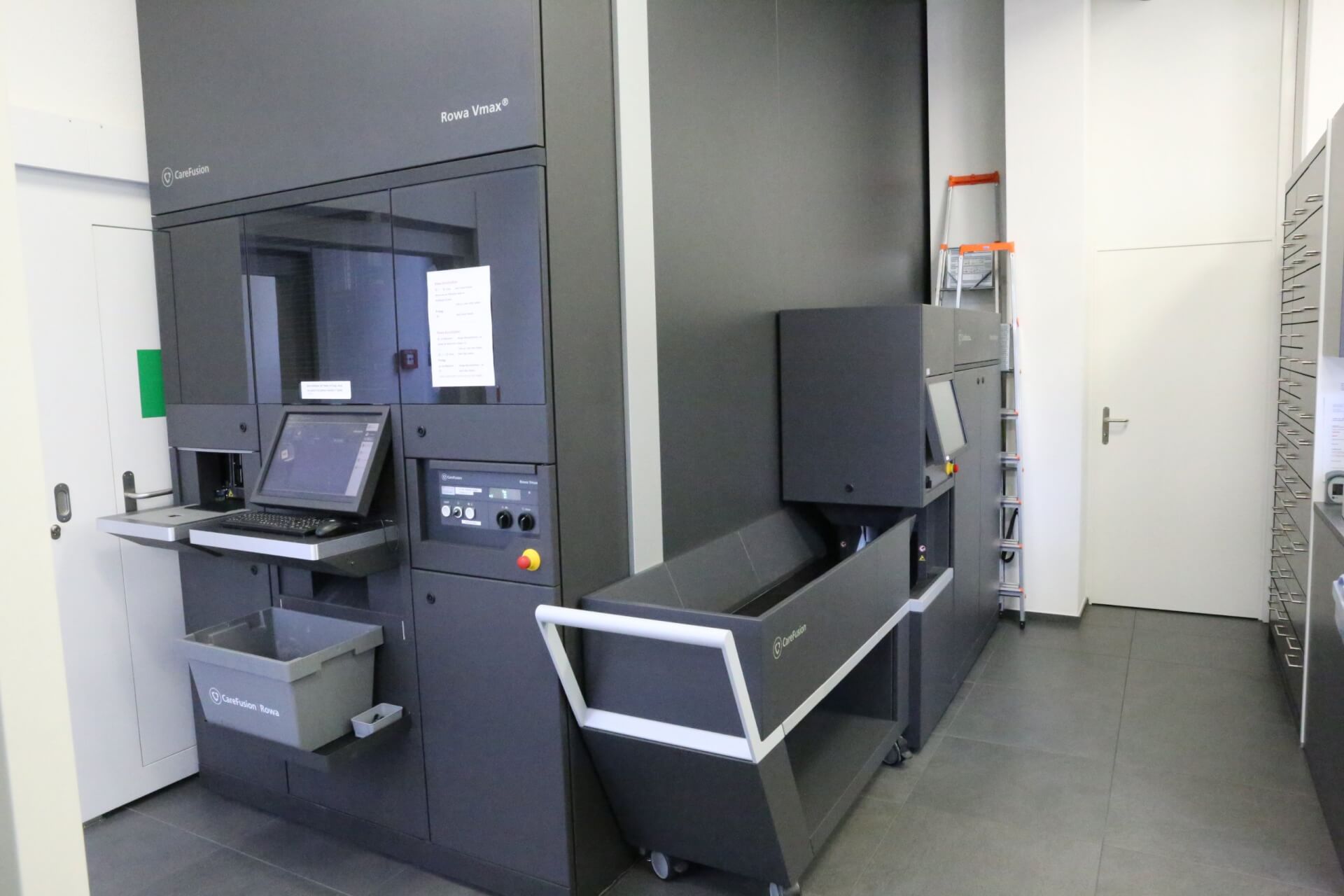 Eine moderne Buchsortiermaschine mit Computerterminal, grauen Beh&auml;ltern und einer Leiter im Hintergrund steht in einem sauberen, weissen Raum des T&auml;fernhofs D&auml;ttwil, der mit effizienter Organisation 15 Jahre Jubil&auml;um feiert.