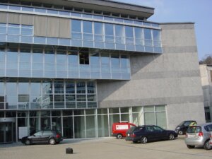 Ein modernes Geb&auml;ude mit grossen Glasfenstern und grauen Steinmauern; davor mehrere parkierte Autos und ein roter Lieferwagen mit einem Koffer auf dem Trottoir - der T&auml;fernhof D&auml;ttwil feiert 15 Jahre Jubil&auml;um.