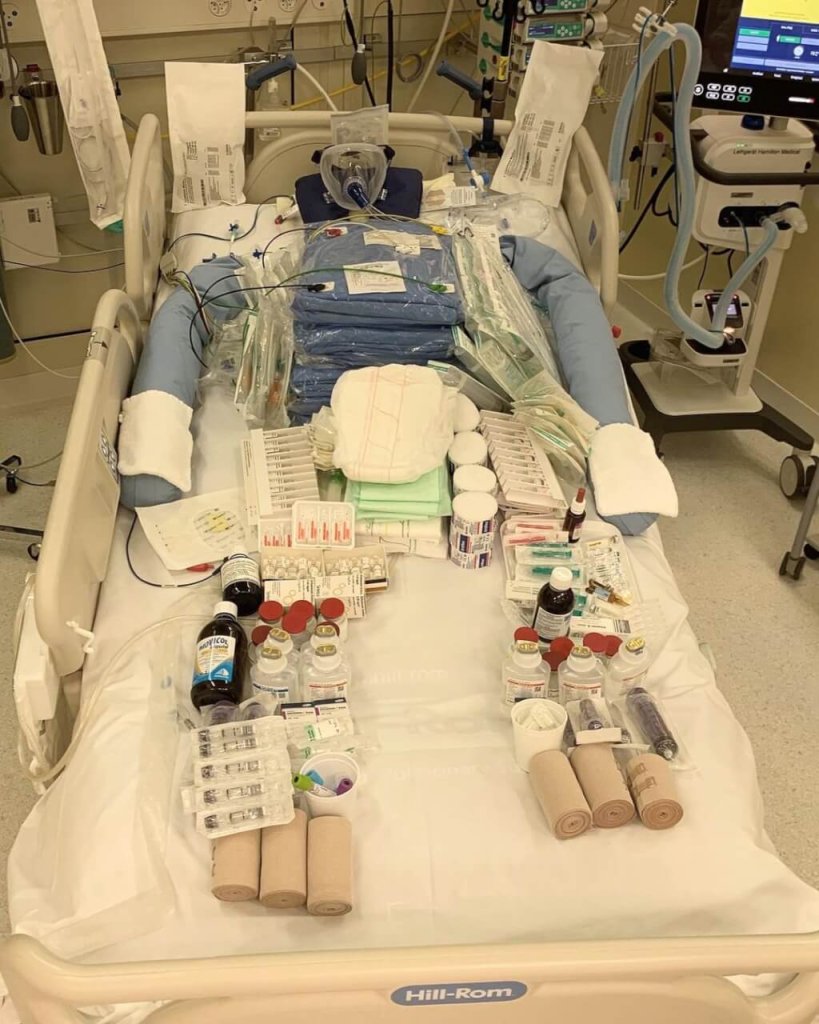 Ein Krankenhausbett, bedeckt mit verschiedenen medizinischen Hilfsmitteln, darunter Verb&auml;nde, Spritzen, Flaschen und COVID-19 Medikamente, in einer klinischen Umgebung mit medizinischen Ger&auml;ten und Monitoren im Hintergrund.