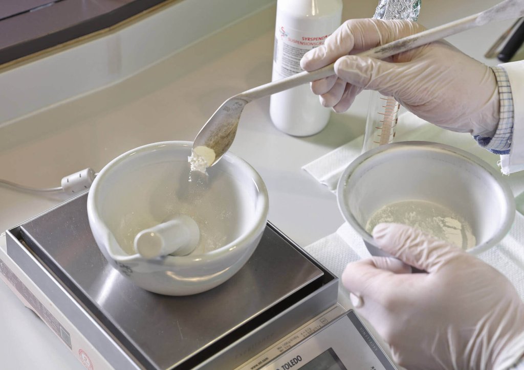 Eine Person mit Handschuhen f&uuml;llt wei&szlig;es Pulver aus einer Schale in einen M&ouml;rser mit einem L&ouml;ffel neben einem St&ouml;&szlig;el auf einer digitalen Waage in einem Labor des &Uuml;berbetrieblichen Kurses (&Uuml;K).