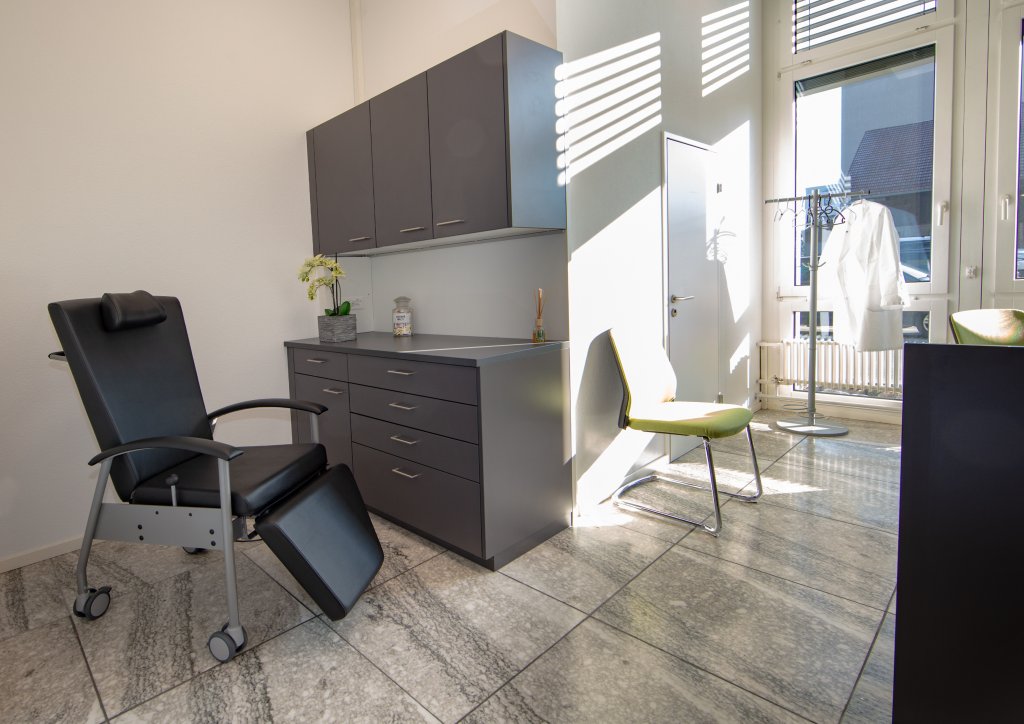 In einem modernen medizinischen Untersuchungsraum am Standort D&auml;ttwil stehen ein schwarzer Lehnstuhl, graue Schr&auml;nke, ein kleiner Tisch mit einer Pflanze, ein gelber Stuhl, ein Laborkittel an einem Haken und Sonnenlicht, das durch grosse Fenster f&auml;llt.