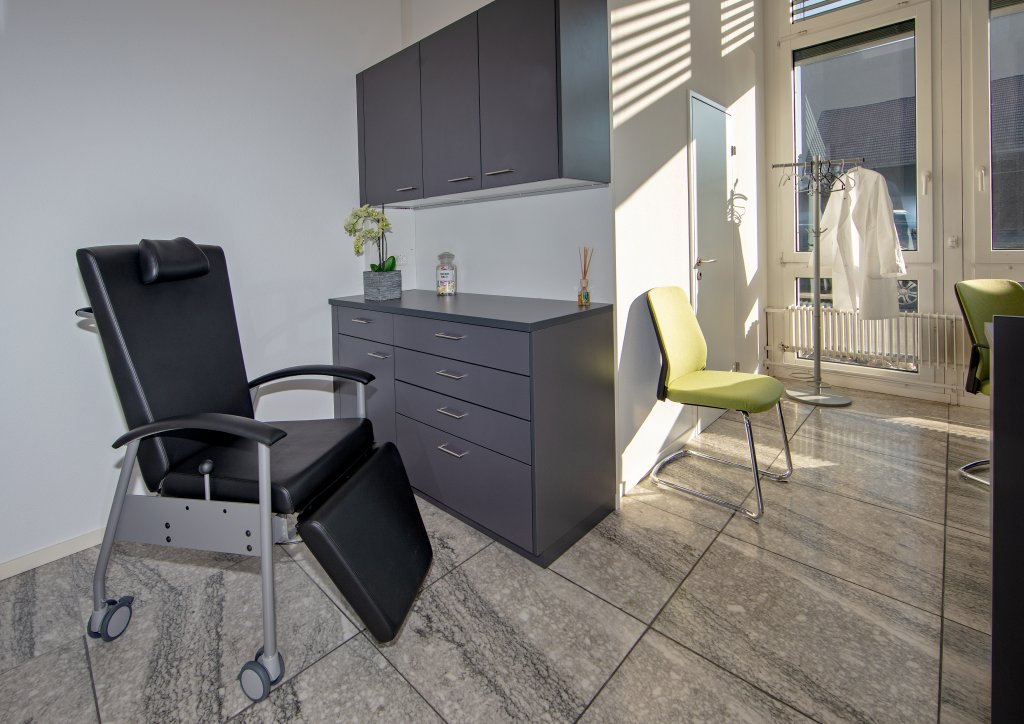 Ein modernes medizinisches Untersuchungszimmer am Standort D&auml;ttwil verf&uuml;gt &uuml;ber einen schwarzen Liegestuhl, graue Schr&auml;nke, einen gr&uuml;nen Stuhl, eine Garderobe mit einem weissen Laborkittel und grosse Fenster, die viel Tageslicht hereinlassen.