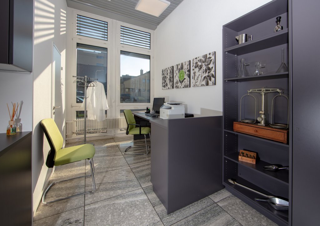 Ein modernes B&uuml;ro am Standort D&auml;ttwil ist mit einem Schreibtisch, gr&uuml;nen St&uuml;hlen, einer Garderobe mit wei&szlig;em Mantel, einem Drucker und Regalen mit wissenschaftlichen Ger&auml;ten wie einer Waage und Glaswaren ausgestattet; gro&szlig;e Fenster sorgen f&uuml;r Tageslicht.